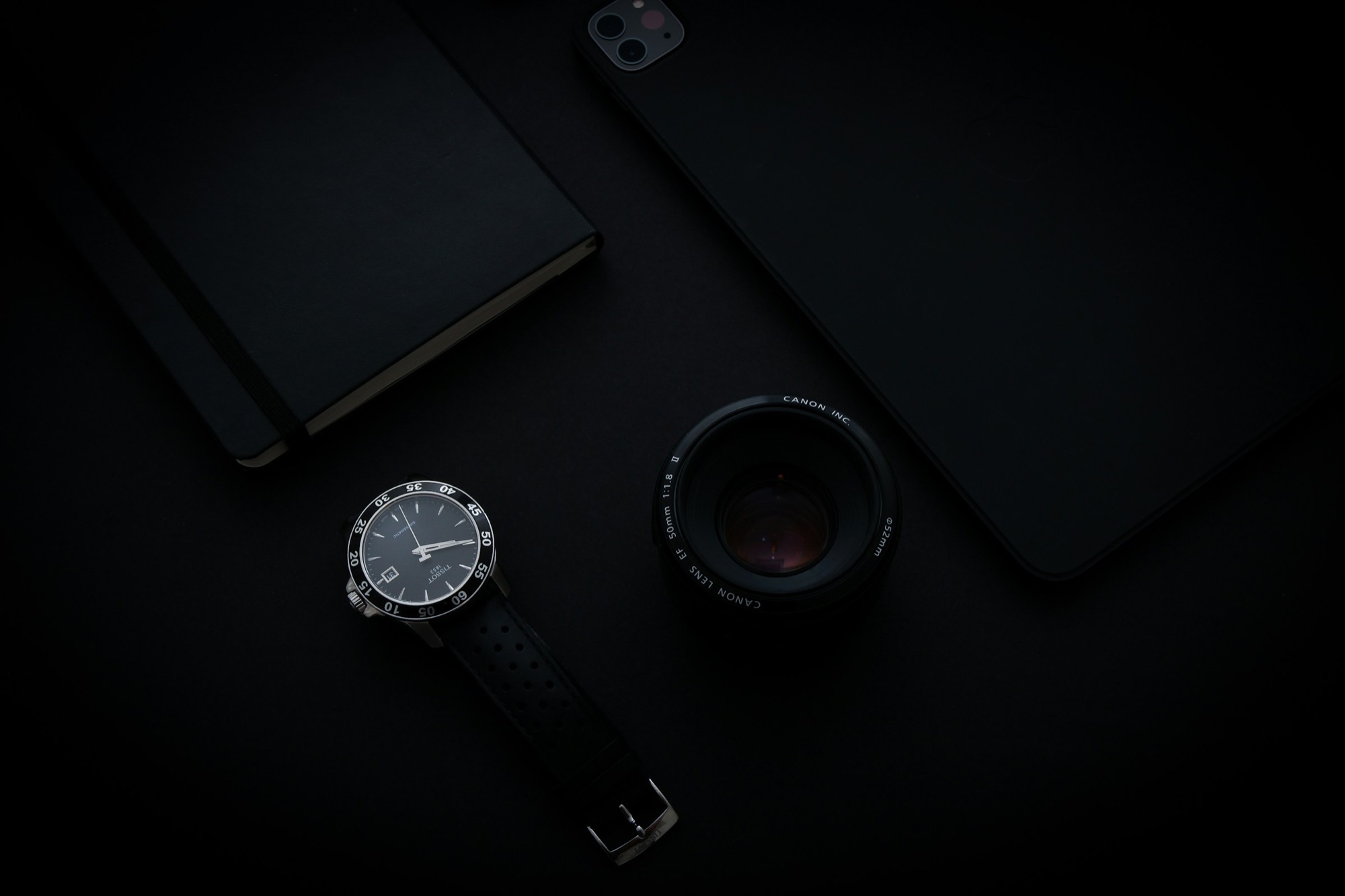 A stainless watch on a perforated leather strap on a dark desk beside a camera lens
