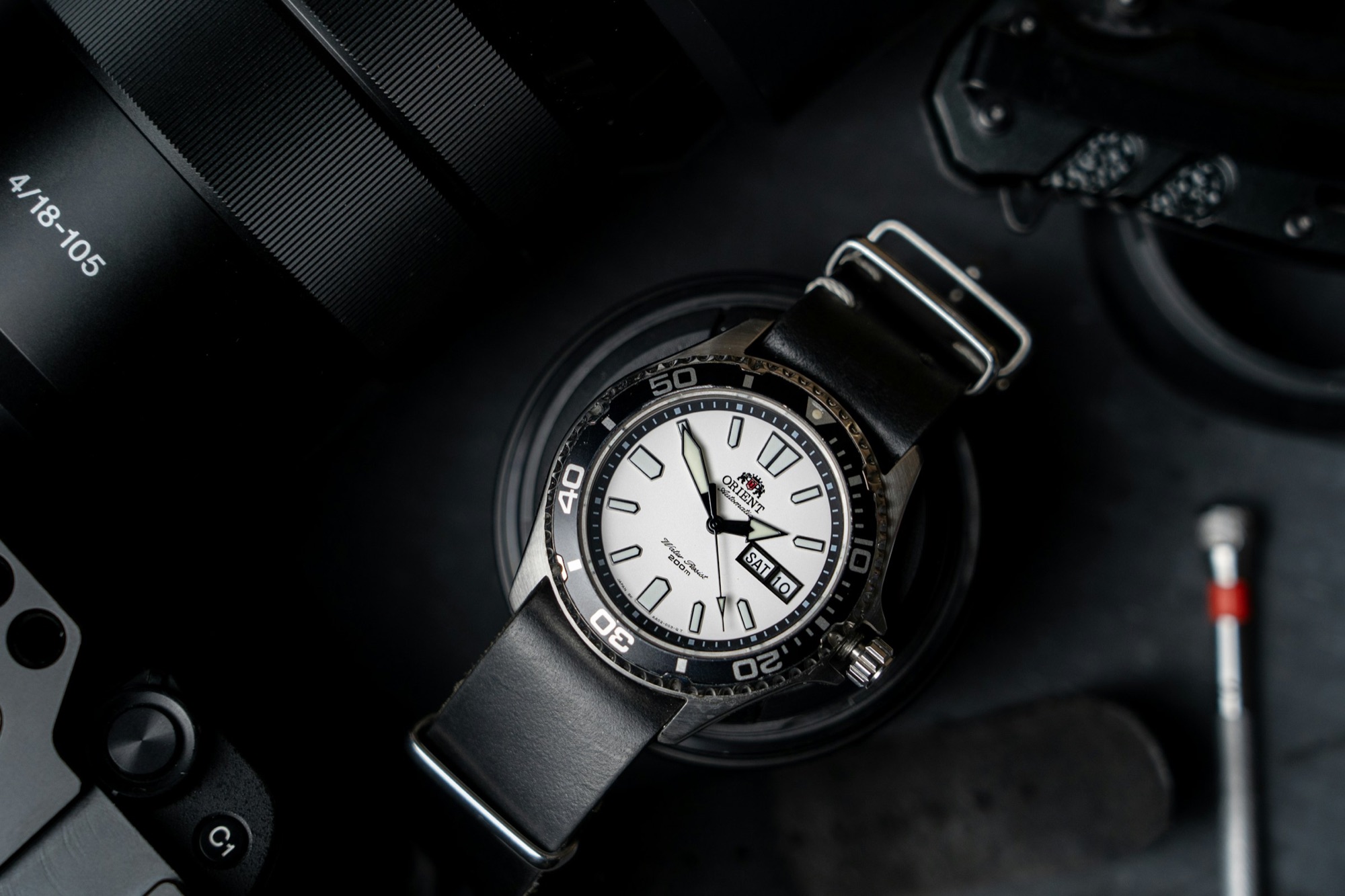A dive watch on a black NATO strap on a dark surface next to a camera and tools